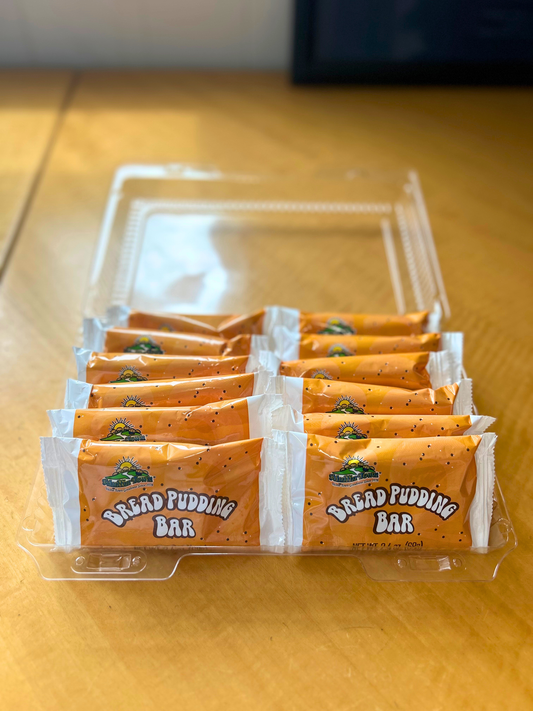 Packaged muffin town bread pudding bars in a clear plastic container on a wooden surface.